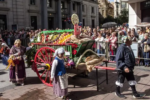 Carro con ofrenda de frutos, pan y verduras, adornado con flores, desfila entre una multitud.