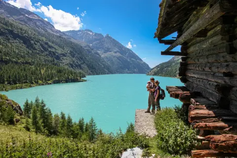 Un couple avec un bébé admirent le lac d'Aoste, entouré de montagnes verdoyantes et d'une cabane en bois rustique.