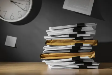 Una pila di documenti su una scrivania, con una nota appiccicata al muro e un orologio che segna il tempo. Chissà ogni quanto si aggiorna il dvr con tutto questo lavoro arretrato.