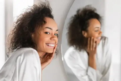 Lächelnde Frau mit lockigem Haar, die sich im Spiegel betrachtet. Ihre Haut strahlt, vielleicht dank Retinol jeden Tag.