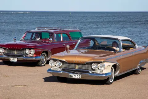 Två klassiska Plymouth Fury 1960, en röd kombi och en guldfärgad coupé, står vid havet.