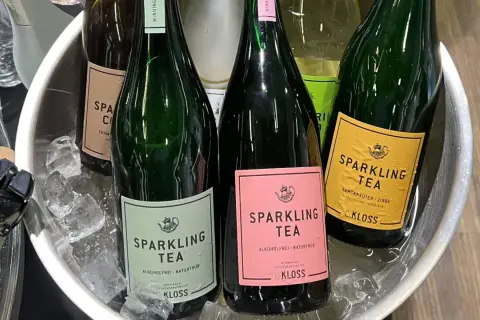Butelki z napojem typu wino cava, schłodzone w lodzie. Etykiety w kolorach zielonym, różowym i żółtym, z napisem "Sparkling Tea".