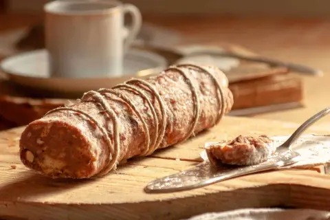 Ein rustikaler salame al cioccolato, gebunden mit Juteschnur, liegt auf einem Holzbrett. Daneben eine Kaffeetasse und ein Stück auf einem Tortenheber.