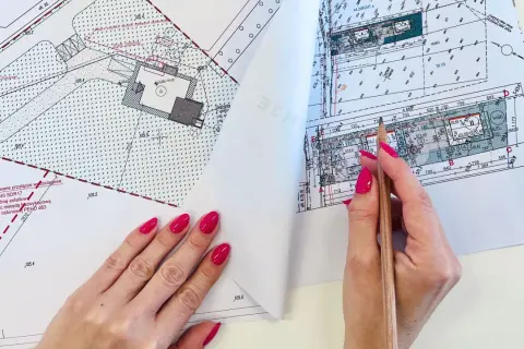 Ręka z różowymi paznokciami wskazuje ołówkiem na mapę do celów projektowych. Widoczne szczegóły działki i budynku.