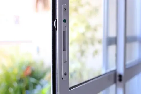Detalle de un mecanismo de cerraduras seguras en una ventana corrediza, con un fondo desenfocado de vegetación y luz.