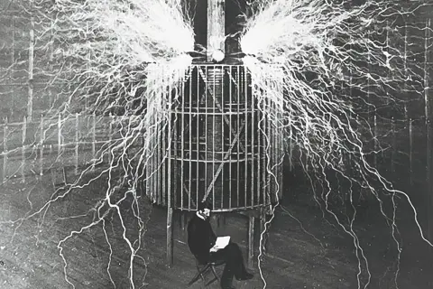 Eksperyment z wyładowaniami elektrycznymi, pokazujący różne rodzaje kondensatorów w akcji. Mężczyzna siedzi spokojnie wśród błyskawic.