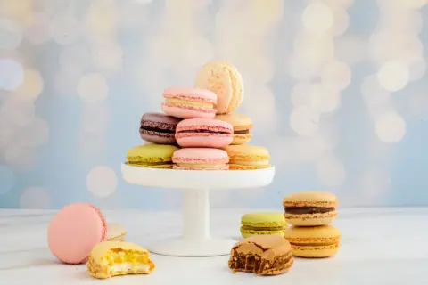 Bunte Macarons auf einem weißen Tortenständer, einige angeschnitten, vor unscharfem Bokeh-Hintergrund.