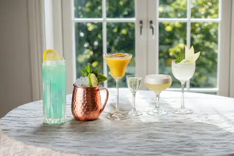 Fem olika vodka drinkar, från en blå long drink till en gyllene passionfruktscocktail och en vit äppeldryck, står uppradade på ett dukat bord.