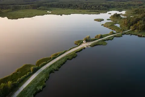 Droga przecinająca jezioro, otoczona zielenią i lasem. W oddali mały domek, jakby cyców na wodzie.