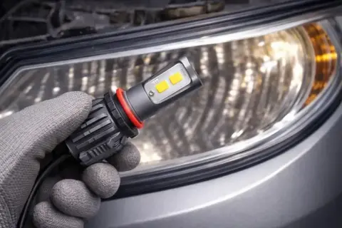 Ręka w rękawicy trzyma nowoczesną żarówkę LED, gotową do wymiany starej lampy halogenowej w reflektorze samochodu.