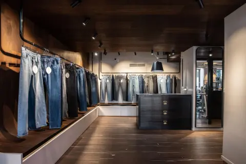 Jeans appesi in un negozio, con una **gamba affusolata** che si nota in primo piano. L'ambiente è moderno, con pavimenti in legno scuro e illuminazione a faretti.