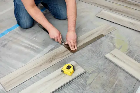 A person is cutting vinyl plank flooring that is not laying flat during installation. A tape measure is nearby.