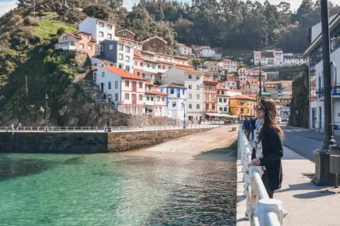 Una mujer admira la costa de **pueblos de Asturias cerca de Galicia**. Casas coloridas se aferran a la ladera verde junto a un mar turquesa.