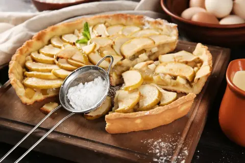 Pyszne ciasta z gruszkami, posypane cukrem pudrem z sitka. Obok dzbanuszek z sosem i miska jajek.