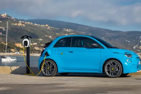 Abarth EV azul cargando junto al mar, con un velero y colinas al fondo.