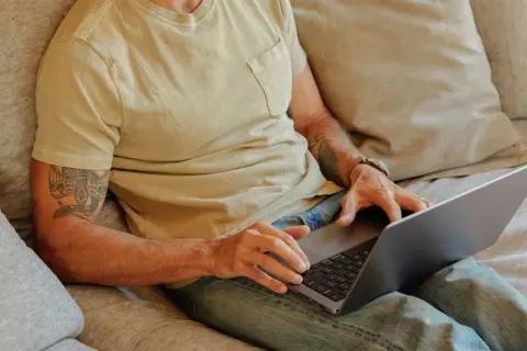 Man med tatuering på armen sitter i soffan med laptop, kanske för att han är hemma under en ny sjukperiod.