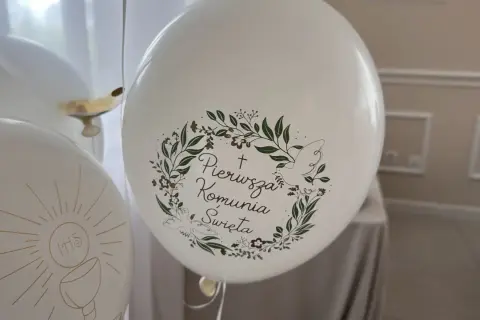 Białe balony z napisem "Pierwsza Komunia Święta" i motywem gołębicy oraz kielicha z hostią.
