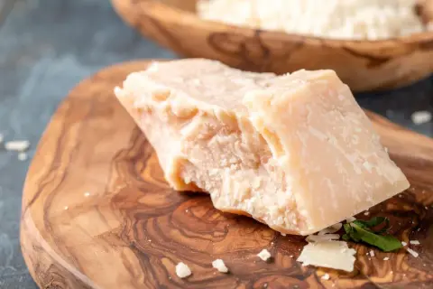 Kawałek sera Grana Padano na drewnianej desce, w tle wiórki sera.