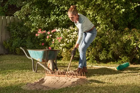 Frau verteilt mit Harke Sand auf dem Rasen. Eine Schubkarre und ein Besen liegen daneben.
