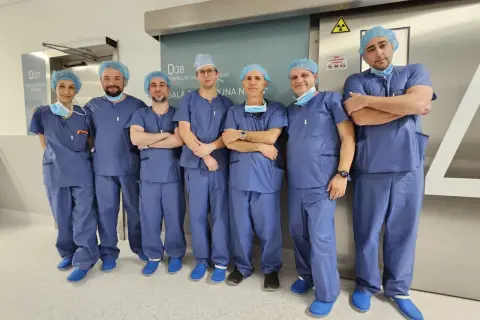 Grupa lekarzy w niebieskich uniformach i czepkach stoi przed drzwiami sali operacyjnej.