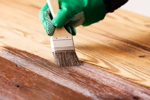 Mano con guante verde aplicando barniz a madera. Proceso para saber como impermeabilizar madera y protegerla.