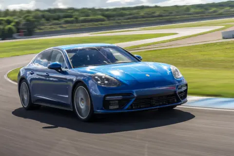 Niebieski Porsche Panamera GTS pokonuje zakręt na torze wyścigowym, z zielonymi wzgórzami w tle.