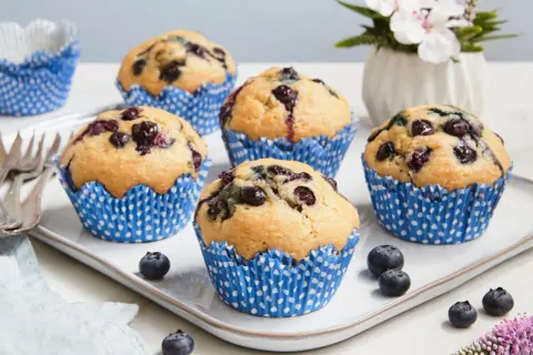 Fünf saftige, vegane Blaubeermuffins in blauen Papierförmchen auf einem weißen Teller. Frische Blaubeeren und eine kleine Blumendekoration runden das Bild ab.