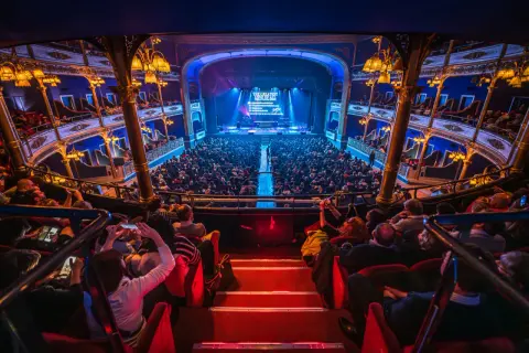 Un pubblico numeroso assiste a uno spettacolo nel Teatro Colosseo di Torino, con luci soffuse e un palco illuminato.