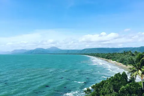 Vacker kustlinje vid Port Douglas med turkost hav, grönskande berg och en gyllene strand under en molnig himmel.