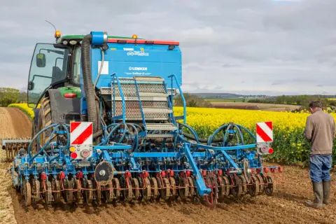 Un tracteur vert et une semoir Lemken Solitair 9 préparent le terrain pour le semis d'avoine de printemps, avec un champ de colza en fleurs en arrière-plan.