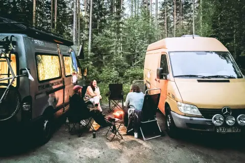 Två husbilar parkerade i skogen, med folk som sitter runt en lägereld. En cykel sitter på takräcket på den ena bilen. En härlig bild av van life.