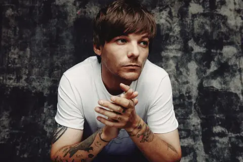 Louis Tomlinson zamyślony, z dłońmi złożonymi przed sobą. Fani czekają na jego nadchodzące wydarzenia.