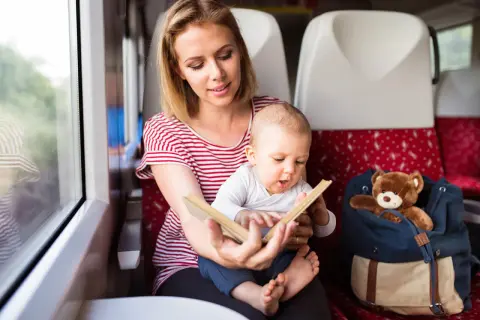 Mama czyta książeczkę rocznemu dziecku w pociągu. Obok siedzi plecak z pluszowym misiem.