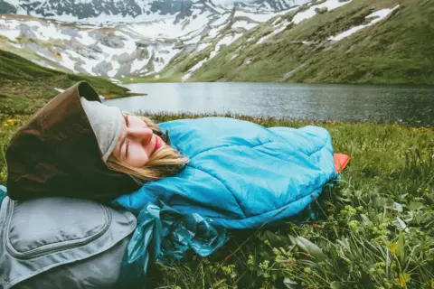 Persona durmiendo en saco azul, con mochila gris, en un prado verde junto a un lago y montañas nevadas. Vivac acampada perfecta.