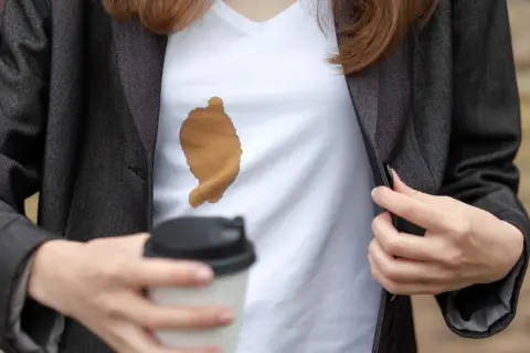 Frau mit Kaffeefleck auf weißem T-Shirt unter dunklem Blazer. Ein Becher Kaffee in der Hand. Tipps, wie man kaffeeflecken aus kleidung entfernen kann.