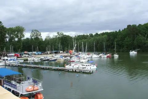 Port w Rucianem Nidzie pełen żaglówek i łodzi. W tle las i pochmurne niebo.