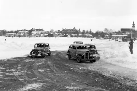 Snöig vinterdag med flera 70 tals bilar redo för race. En åskådare står vid sidan av banan.
