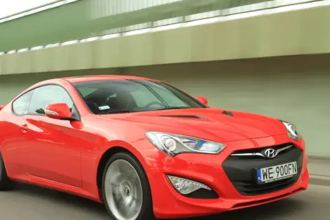Czerwony Hyundai Genesis Coupe 3.8 V6 pędzi pod zielonym wiaduktem.