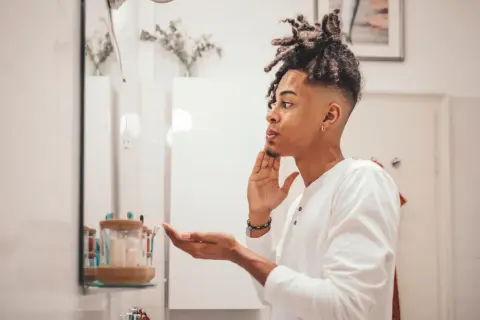 Mann mit Dreadlocks im Spiegel, der sich die Haut richtig reinigen möchte.