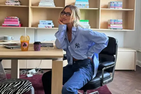 Chica con gafas sentada en una silla de oficina, luciendo un outfit oficina verano hombre: camisa azul oversize y vaqueros.
