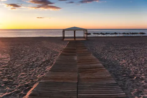 Pasarela de madera hacia una pérgola en la playa de Puerto Rey al atardecer. Sombrillas alineadas al fondo.