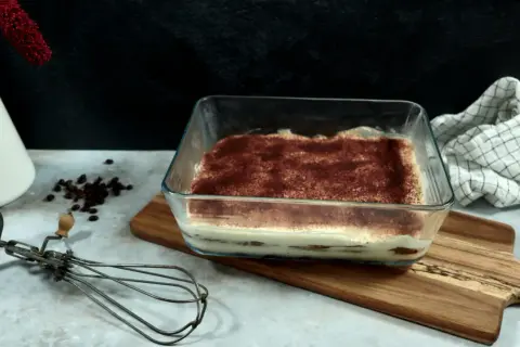 Un délicieux tiramisu 6 personnes, prêt à être dégusté, saupoudré de cacao. Un fouet ancien et des grains de café complètent la scène.