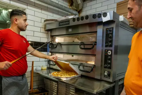 Två pizzabagare jobbar vid en stor pizzaugn. En tar ut en färdig pizza i Sverige.