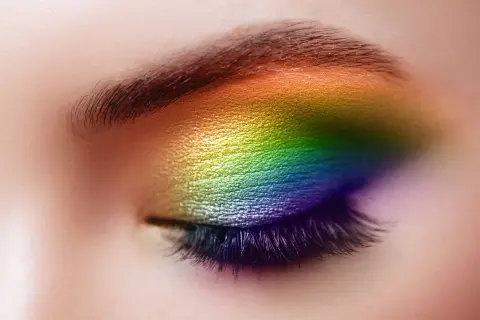 Un œil fermé orné d'un maquillage arc-en-ciel vibrant, du jaune au violet, sublimant le regard. Un maquillage yeux bleus audacieux et coloré.