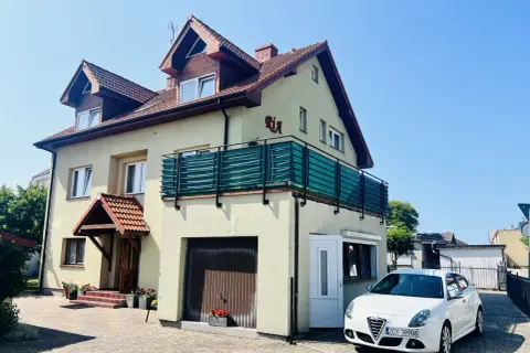 Tanie kwatery prywatne nad morzem. Dom z balkonem i białym samochodem przed posesją, otoczony zielenią.