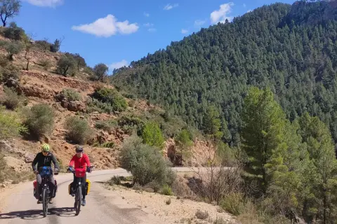 Dos ciclistas exploran rutas gravel por un camino rural rodeado de montañas boscosas y terreno rocoso bajo un cielo azul.