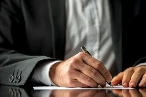 Mężczyzna w garniturze pisze piórem na białej kartce papieru.