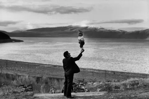 Un homme soulève un bébé au-dessus de l'eau, une scène qui semble être une mise en scène poétique avec des montagnes en arrière-plan.