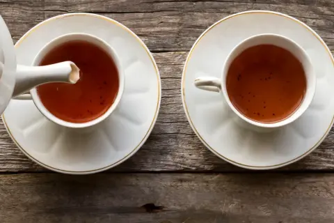 Dwie filiżanki z herbatą pu erh, z których jedna jest nalewana z czajniczka. Napój ma głęboki, bursztynowy kolor, a jego właściwości zdrowotne są cenione od wieków.