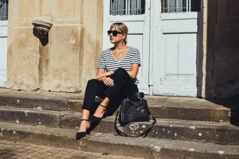 Elegancka kobieta w czarnych cygaretkach i bluzce w paski, siedząca na schodach. Stylowa torebka Michael Kors dopełnia całość.